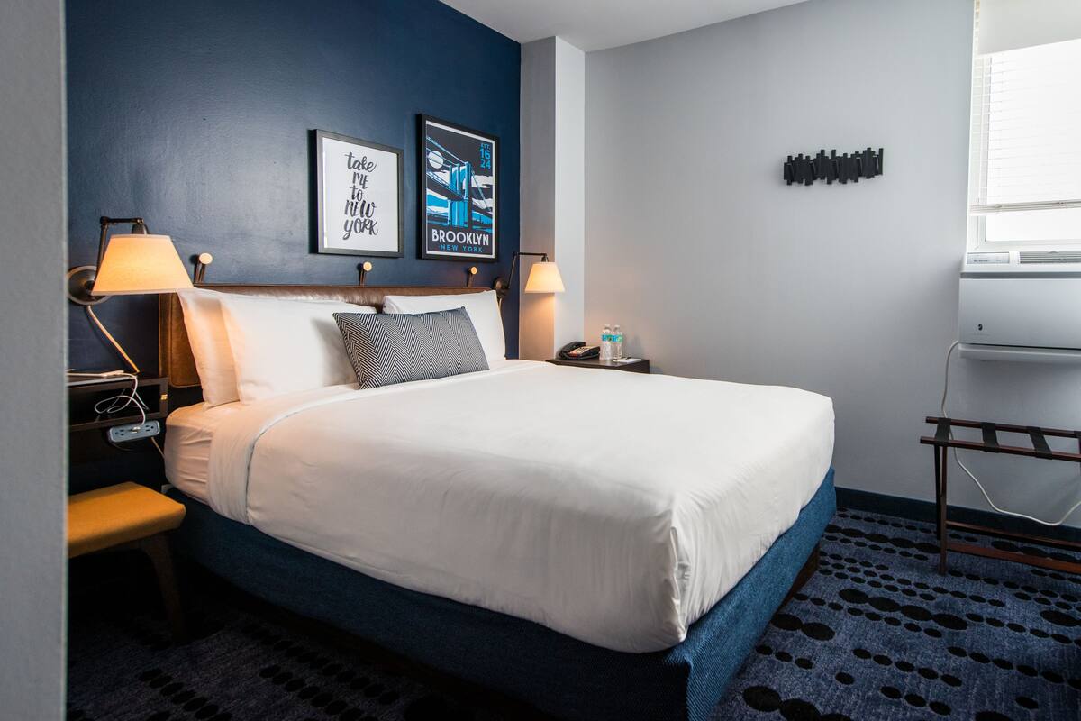 Une chambre d’hôtel avec un lit, deux lampes fixées au mur, un tableau encadré, une petite table avec un téléphone et des bouteilles d’eau, et une fenêtre avec des stores.