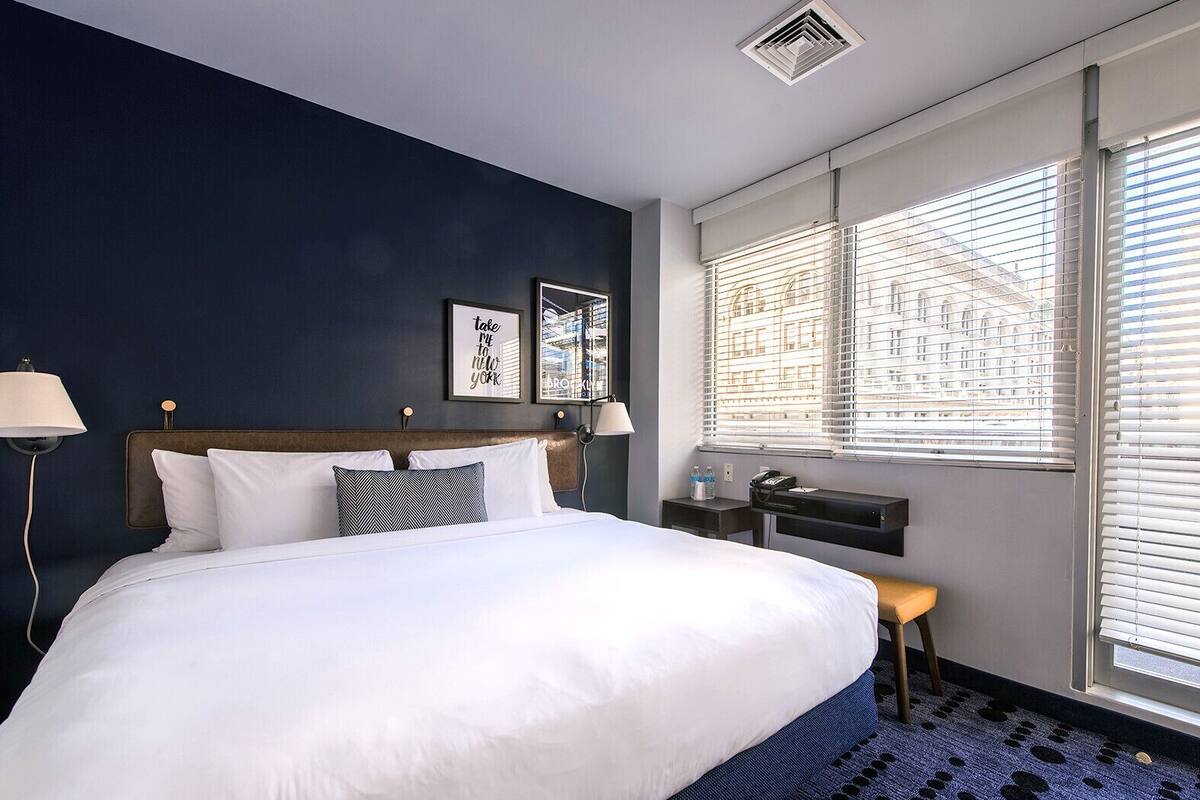 Une chambre d’hôtel avec un grand lit, un bureau avec une chaise, une fenêtre avec des stores et une bouche d’aération fixée au mur.