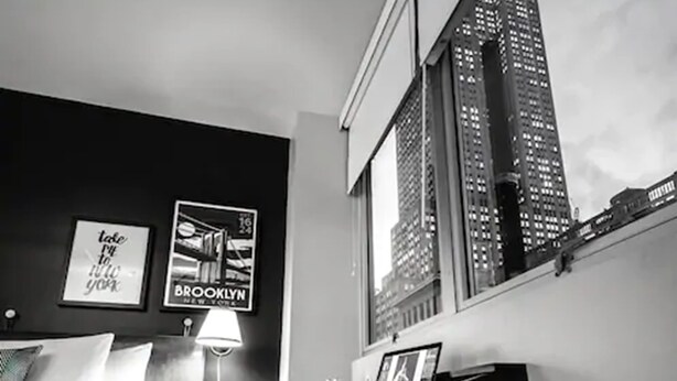 A hotel room with a bed, a desk, a lamp, and a window overlooking a cityscape.