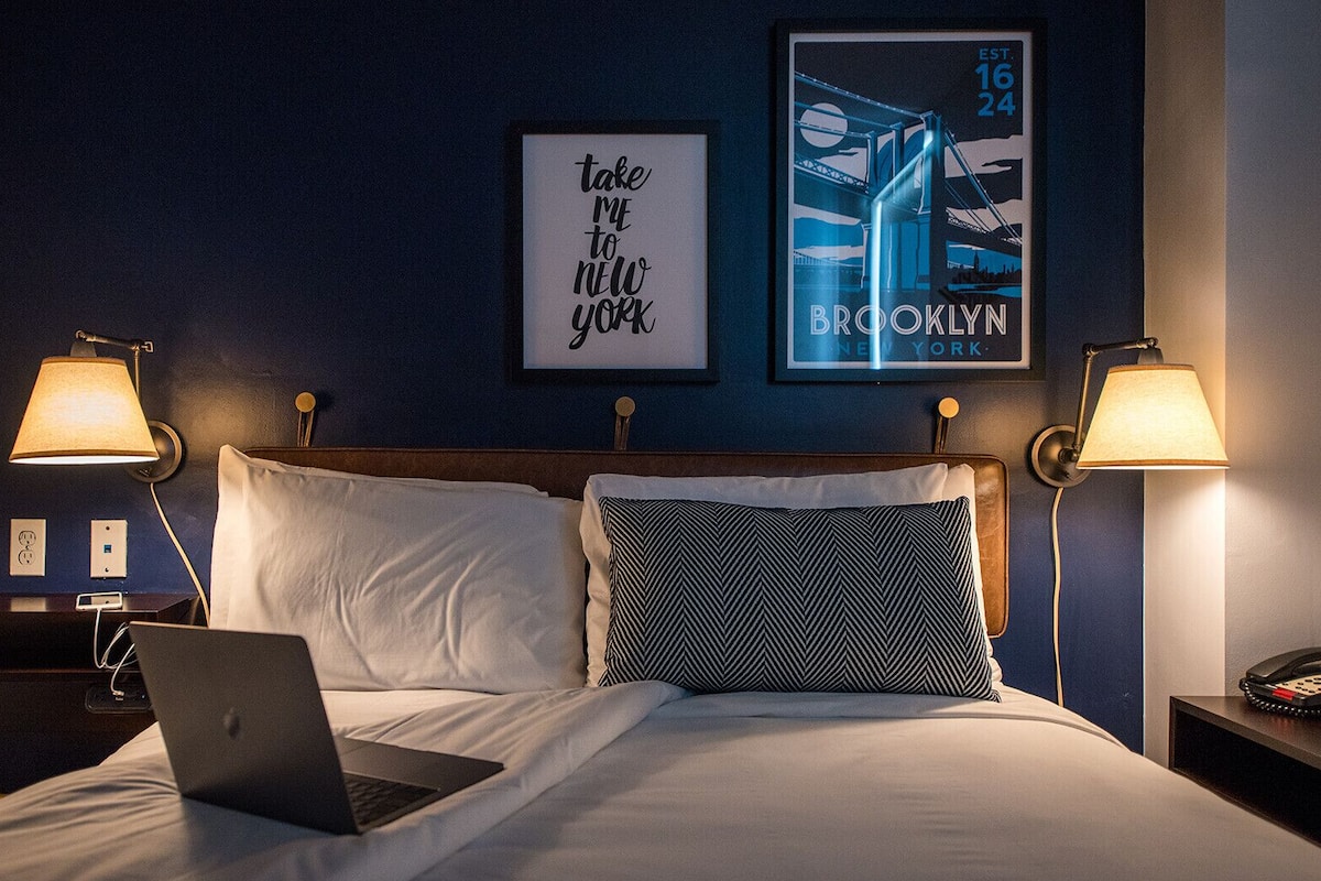 Une chambre d’hôtel avec un lit, deux lampes de chevet, un ordinateur portable et deux tableaux encadrés au mur.