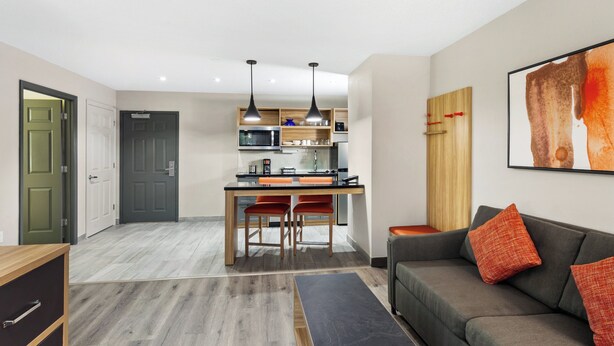 A modern kitchen with a dining area, a grey sofa with orange cushions, and a wooden cabinet.