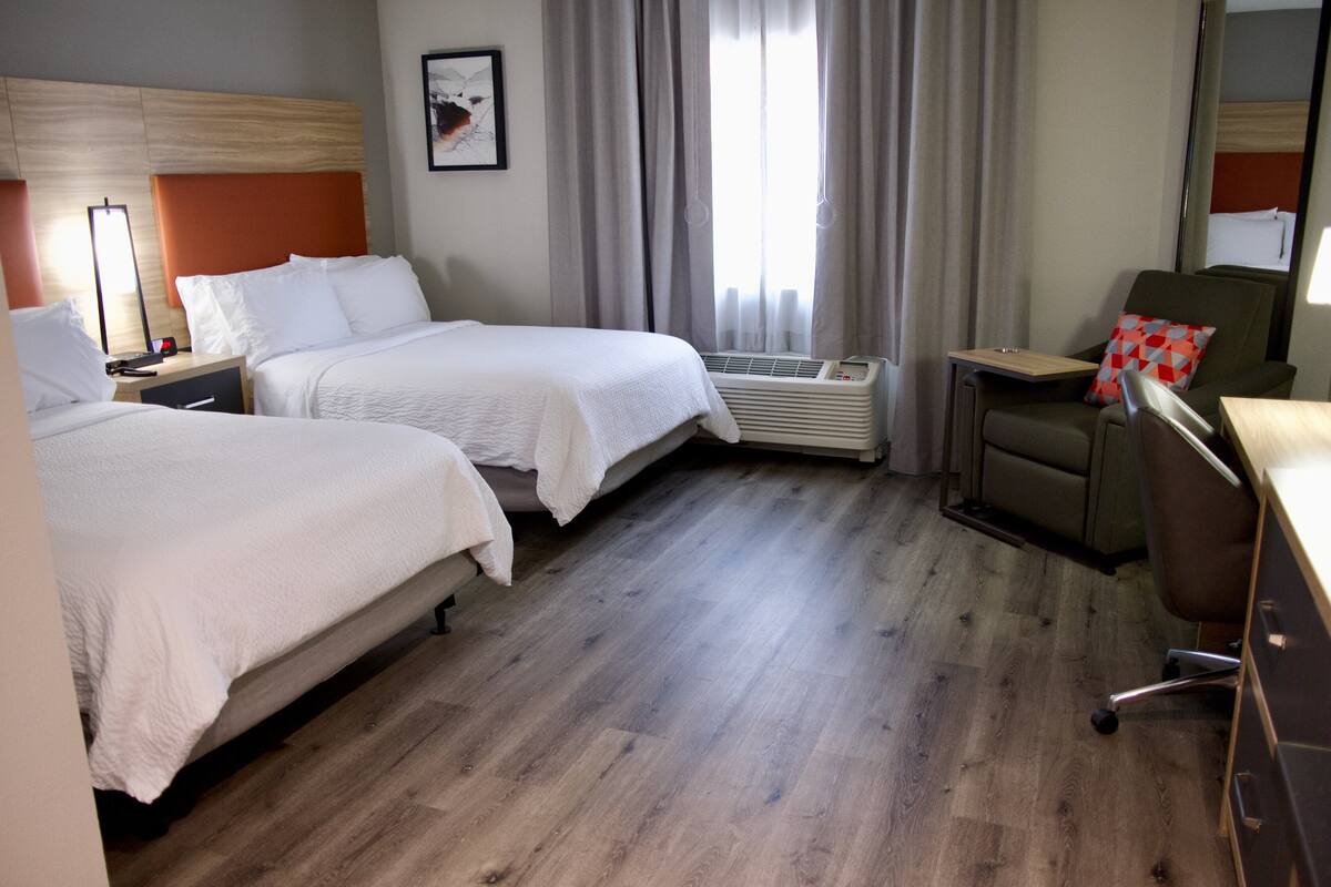 A hotel room with two beds, a desk, a chair, a heater, and a window with curtains.