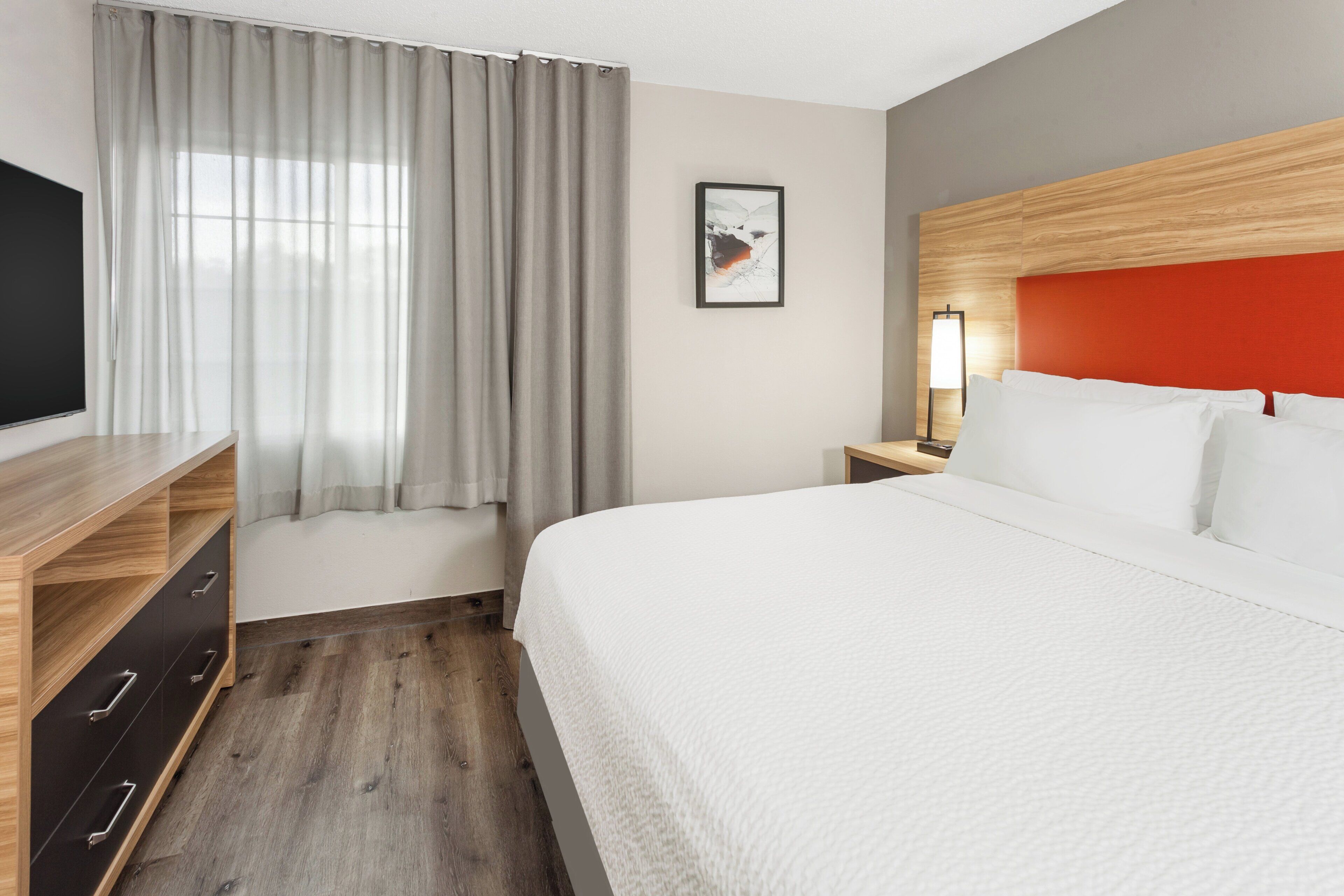A hotel room with a bed, a TV, a nightstand with a lamp, a wooden dresser, and a window with curtains.