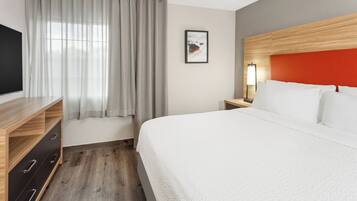 A hotel room with a bed, a TV, a nightstand with a lamp, a wooden dresser, and a window with curtains.