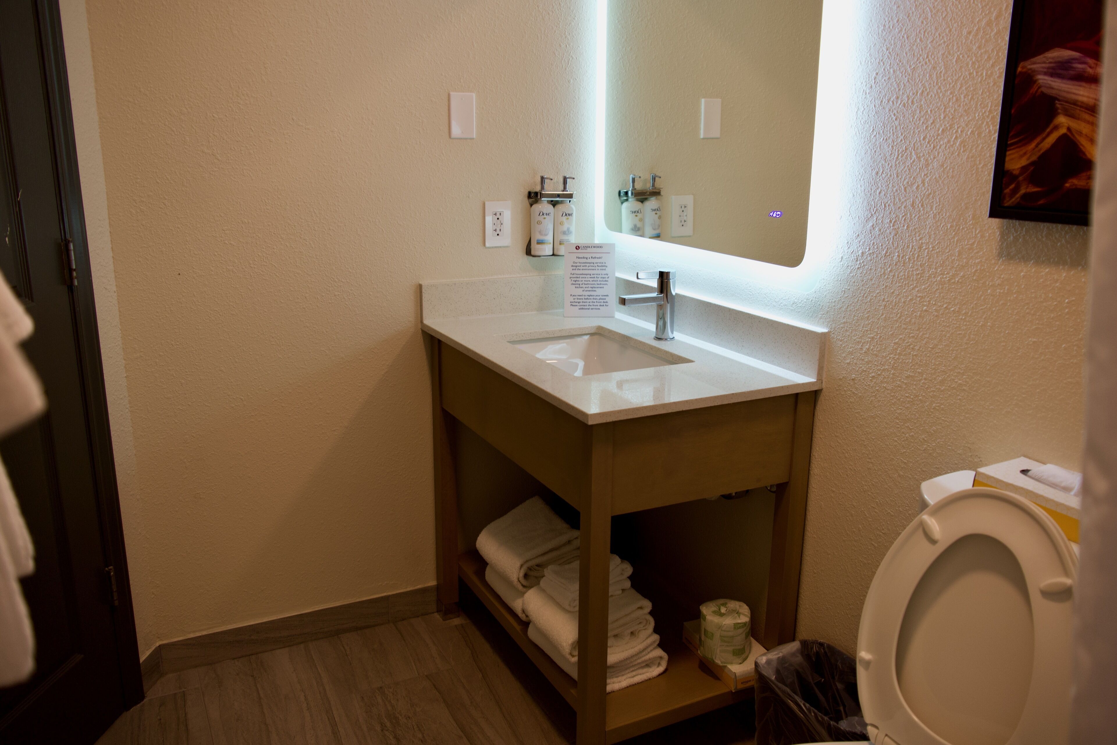 Een badkamer met een witte wastafel, spiegel en toilet. Handdoeken en een vuilnisbak zijn zichtbaar.