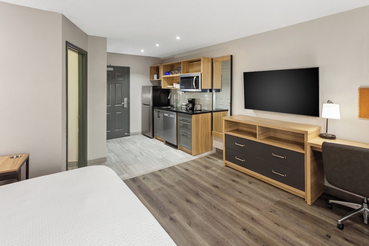Una habitación de hotel moderna con una cama grande, un escritorio con silla, un televisor de pantalla plana, una kitchenette y un baño.