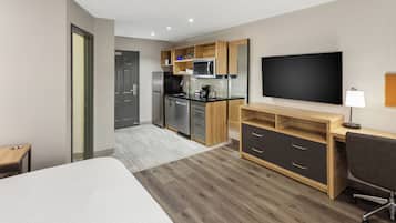 A modern hotel room with a large bed, a desk with a chair, a flat-screen TV, a kitchenette, and a bathroom.
