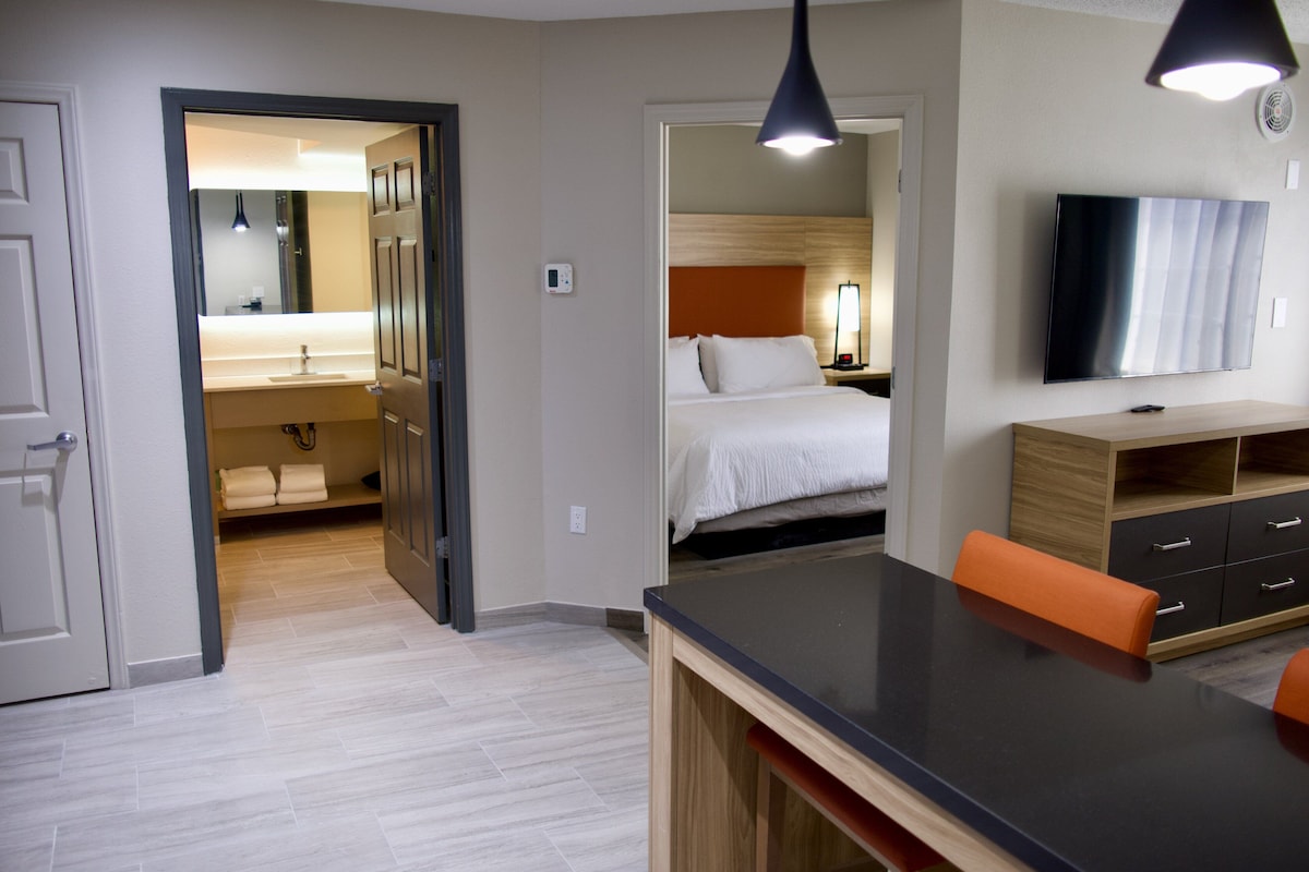 Habitación de hotel con cama, televisión, escritorio con silla y baño visible a través de una puerta abierta.