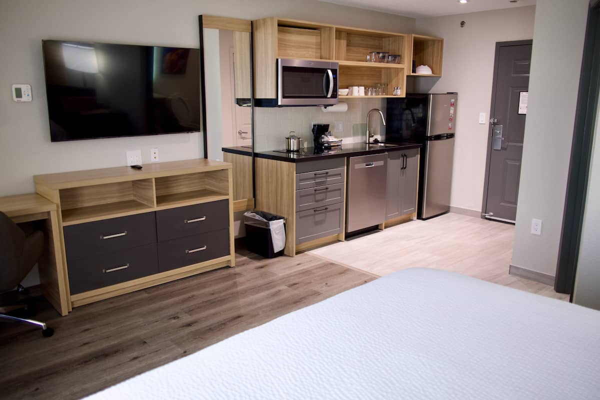 A compact hotel room with a kitchenette, a bed, a desk, and a TV.