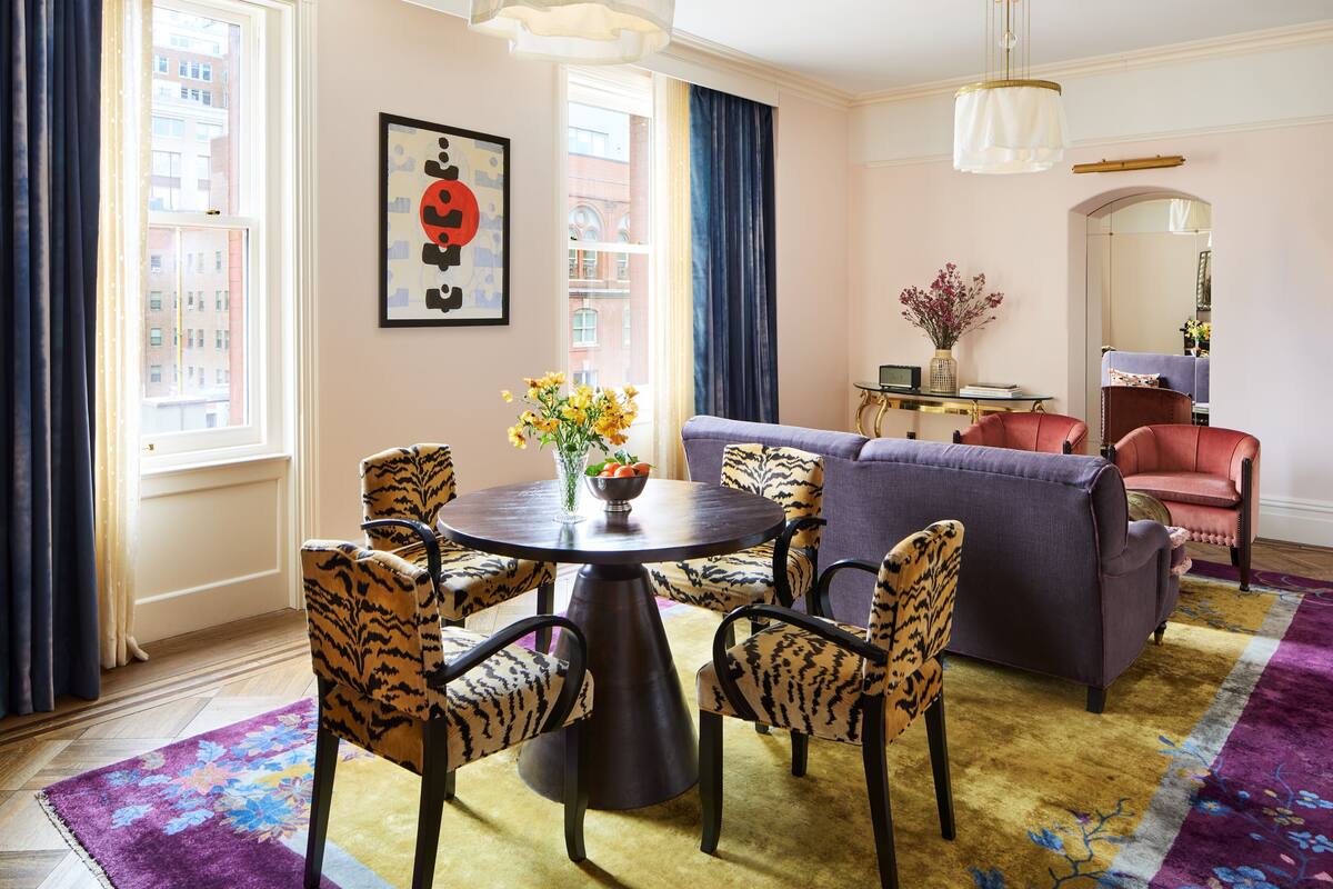 A living room with a round table, two leopard print chairs, a purple sofa, and a red armchair. There is a vase with flowers on the table, and a painting on the wall.