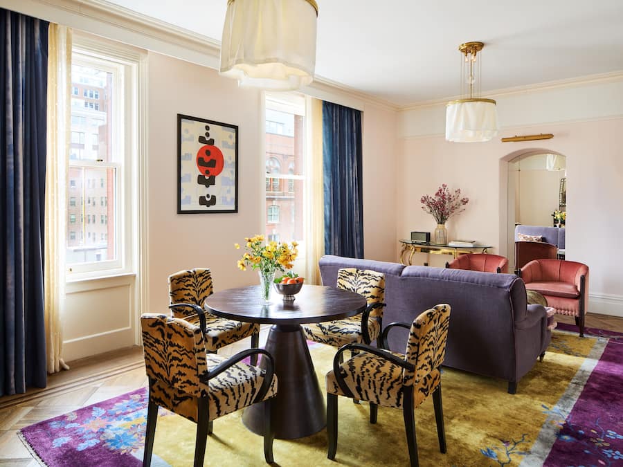 Una sala de estar con una mesa redonda, dos sillas estampadas de leopardo, un sofá morado y un sillón rojo. Sobre la mesa hay un florero con flores, y en la pared, un cuadro.