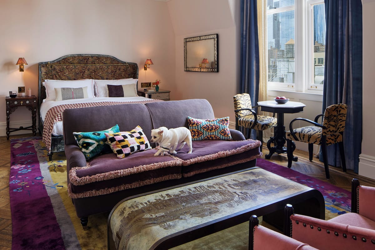 A hotel room with a large bed, a sofa, a small table, and a view of the city through a window.
