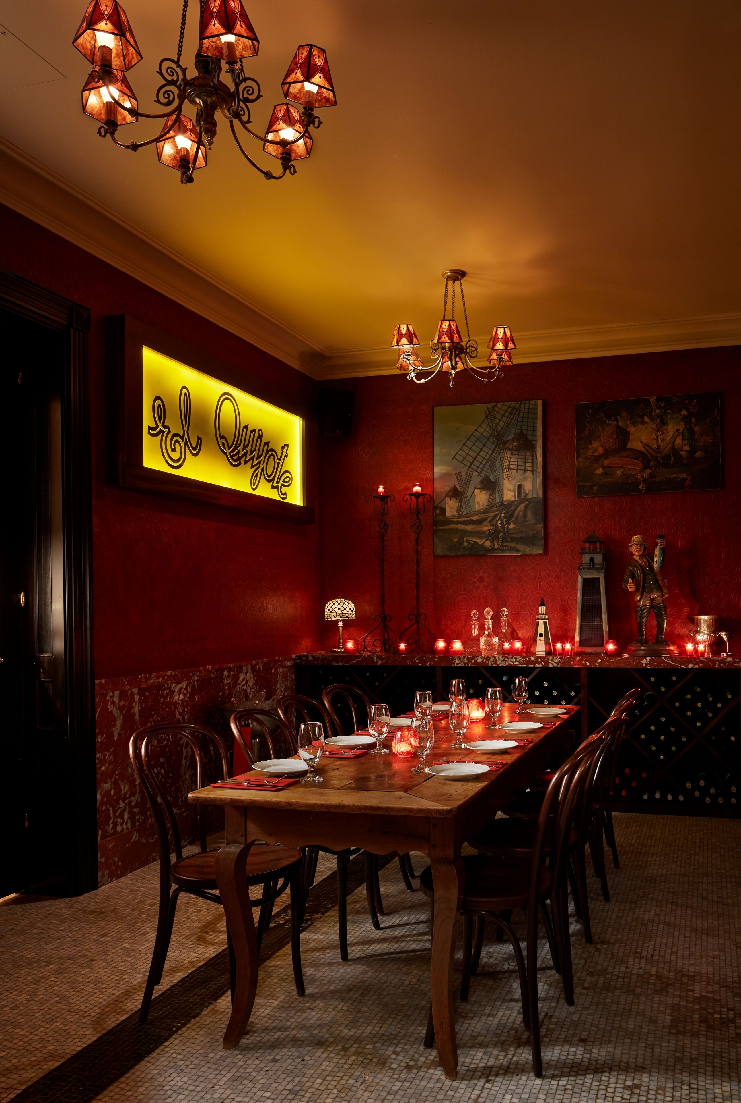 A dining area with a long wooden table set for six, surrounded by chairs. A neon sign above the table reads "El Quijote."