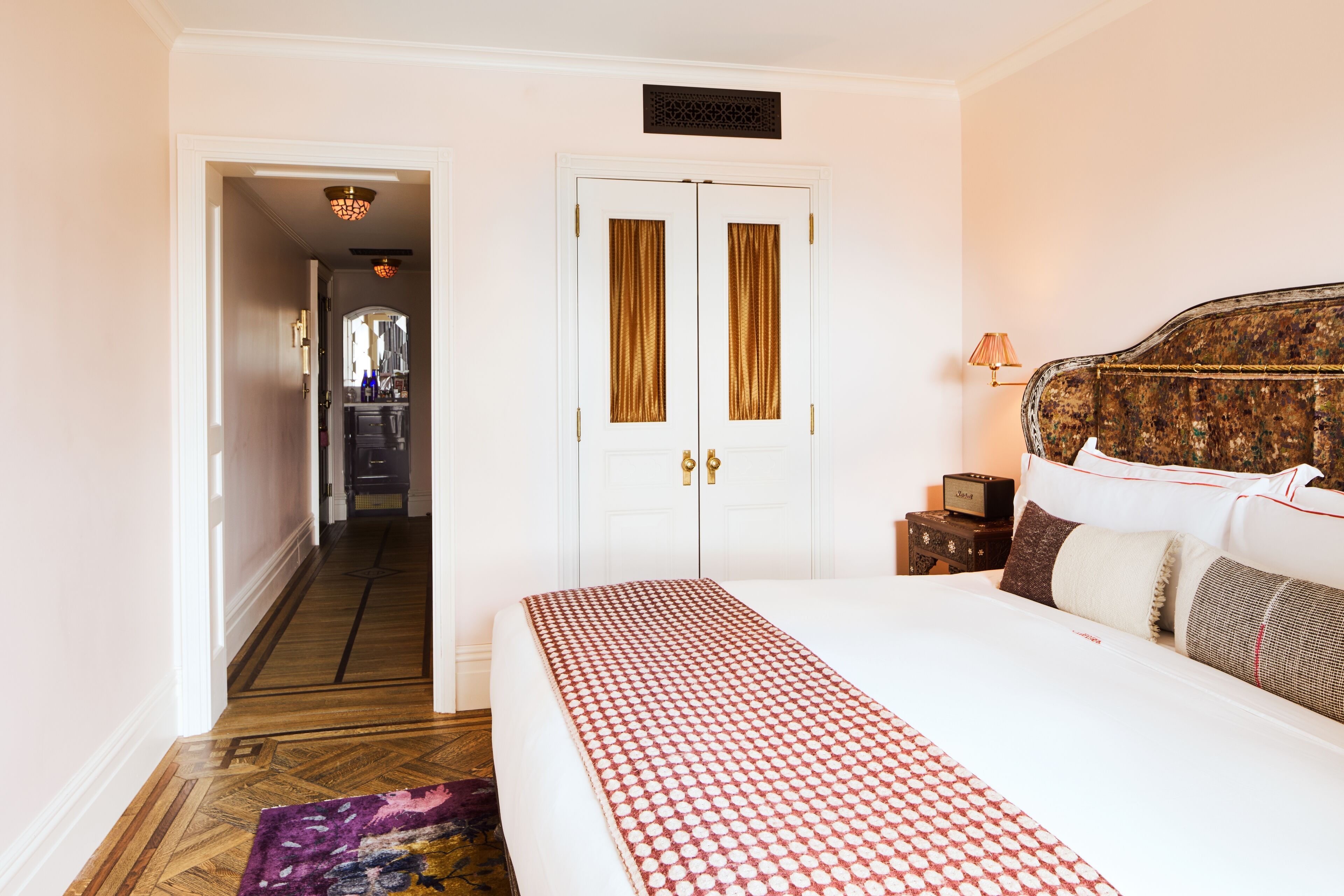 A hotel room with a large bed, a wardrobe with two doors, a nightstand with a lamp, and a view of the hallway.