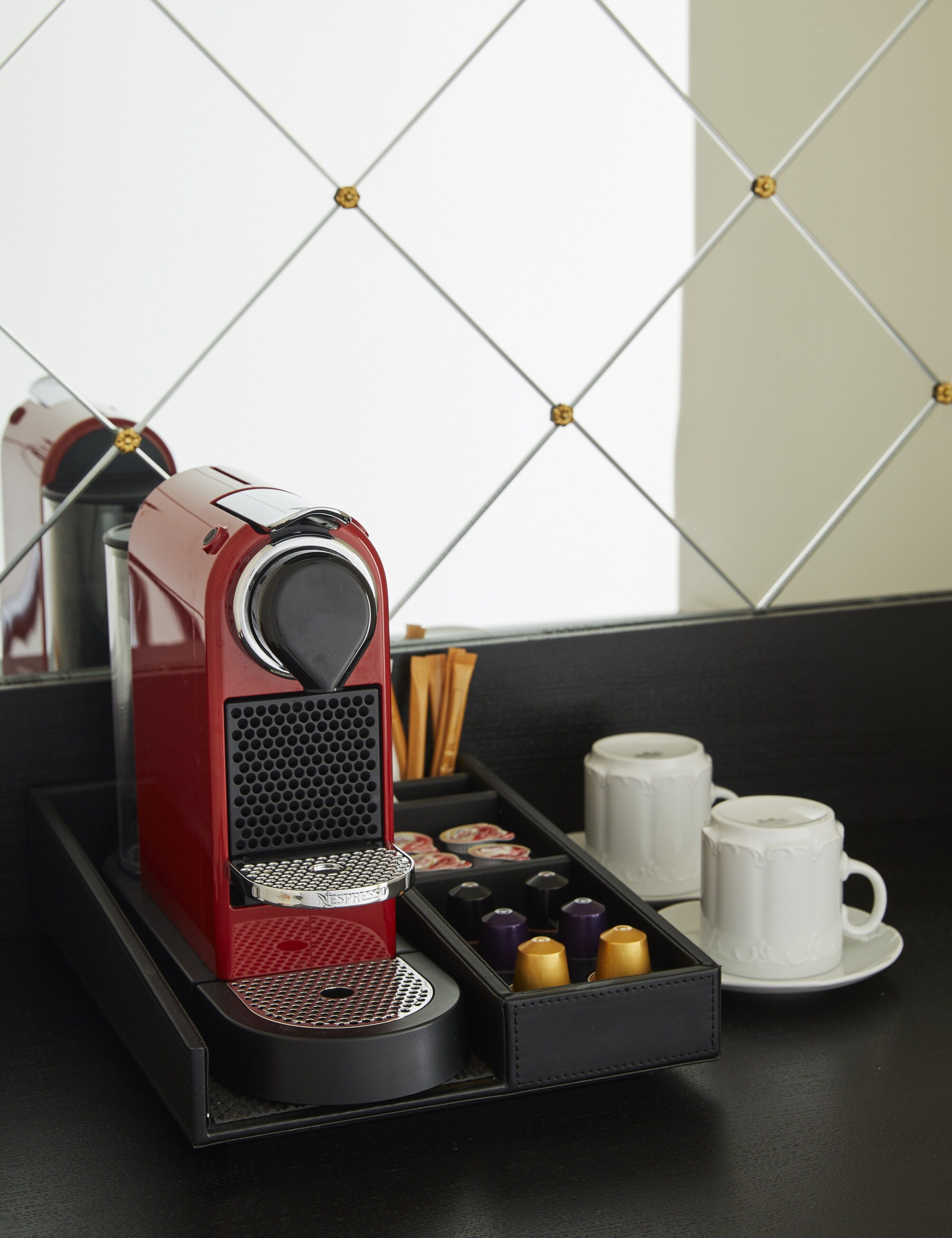 A red espresso machine with a tray of coffee pods and two white cups on a black tray.