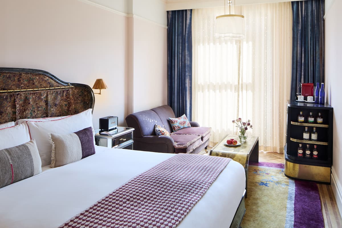 A hotel room with a large bed, a desk, a chair, a minibar, and a window with curtains.