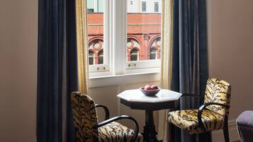 A corner room with a large window, two patterned chairs, a small table with a bowl, and a bed with a patterned blanket.