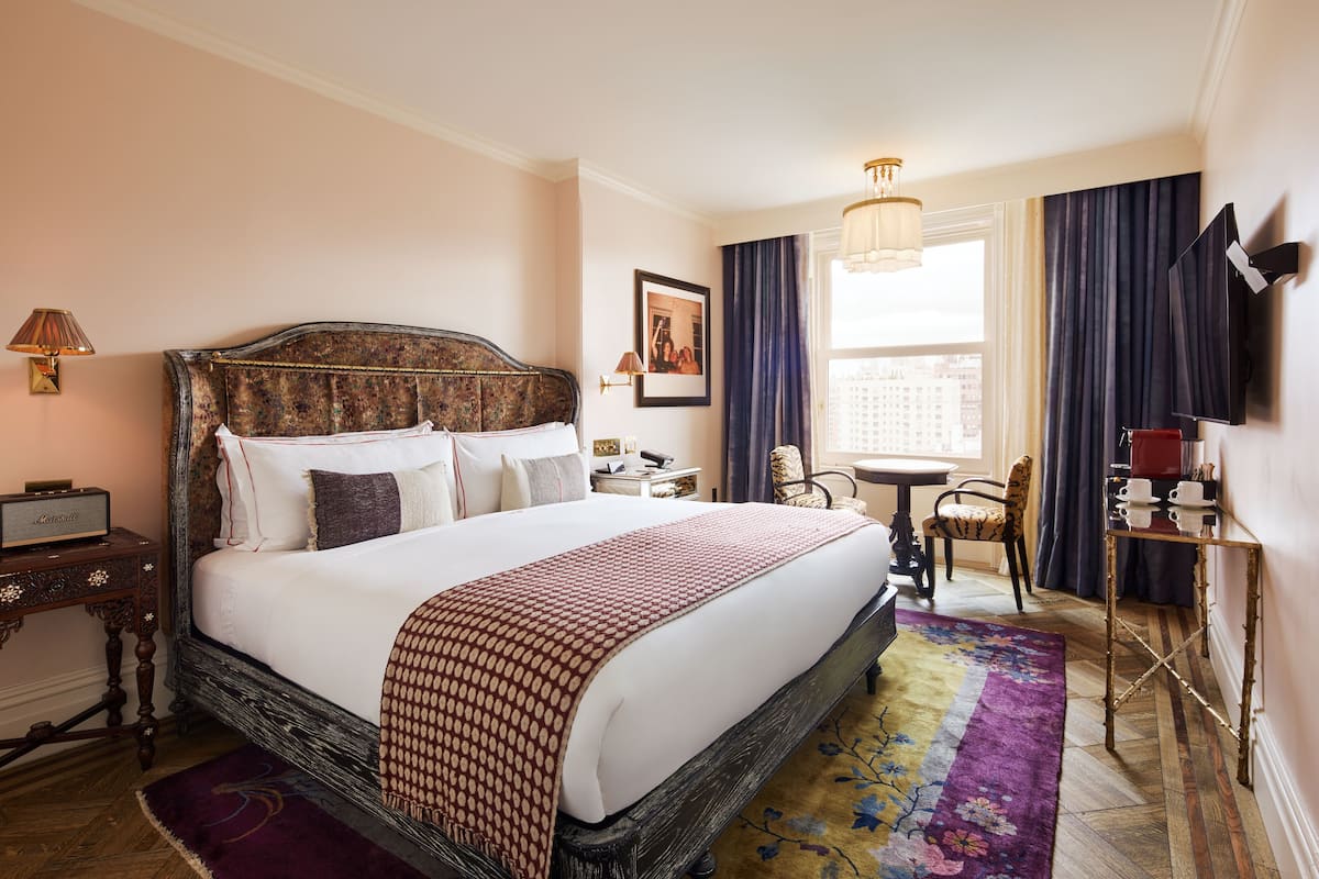 A neatly arranged hotel room with a large bed, a bedside table with a lamp, a small table with a chair, and a TV mounted on the wall.