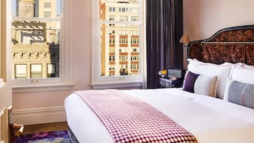A hotel room with a large bed, a view of city buildings, a chandelier, and a nightstand with a lamp.