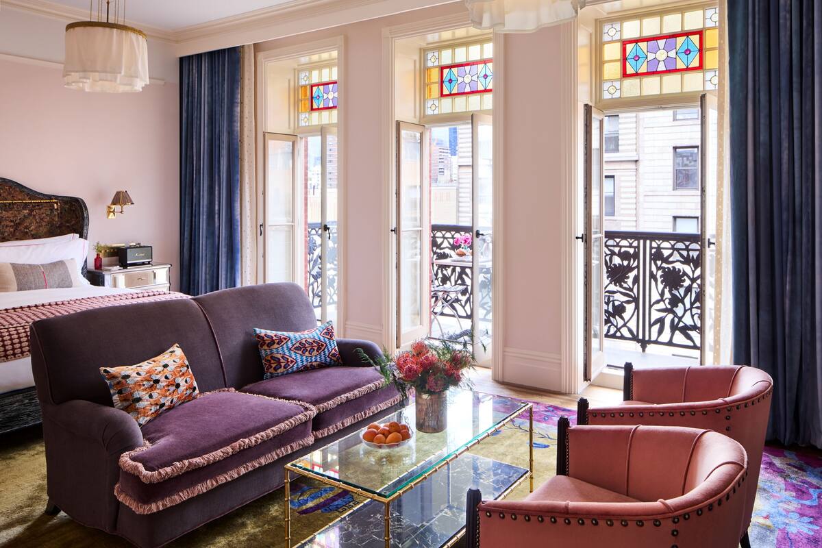 A hotel room with a bed, purple sofa, armchair, and a glass coffee table.