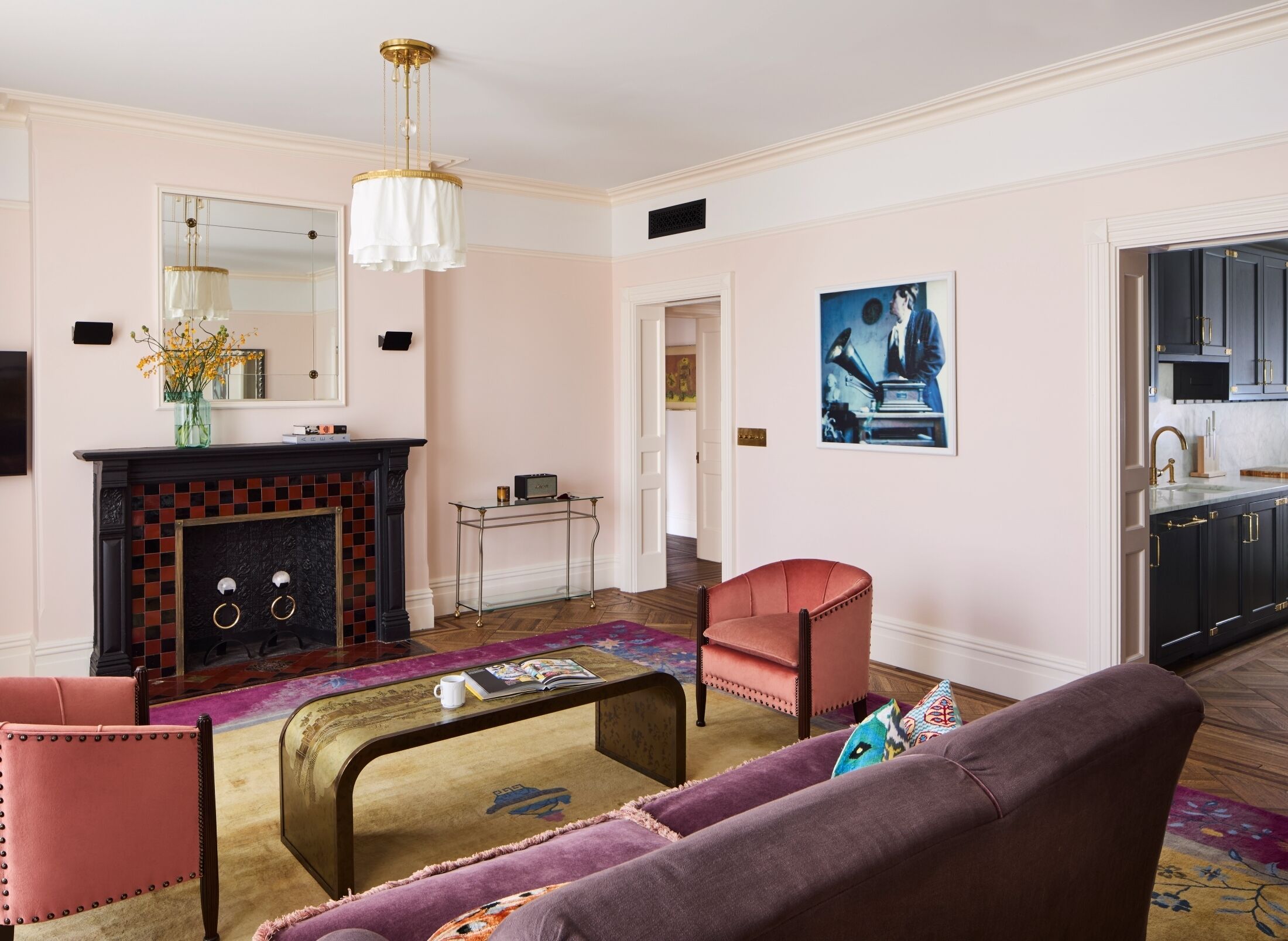 A living room with a fireplace, a coffee table, a sofa, and a chair. There is a kitchen visible through an open doorway.