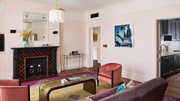 A living room with a fireplace, a coffee table, a sofa, and a chair. There is a kitchen visible through an open doorway.