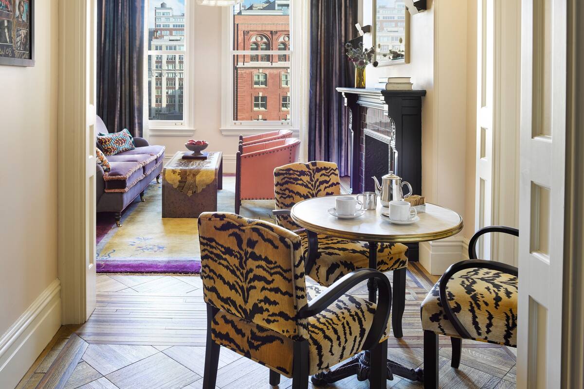 A hotel room with a dining area, a sofa set, a fireplace, and a view of the cityscape.