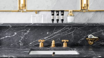 A bathroom with a marble countertop, a sink, and three gold faucets. There are also three black tubes with white labels and a small glass jar.