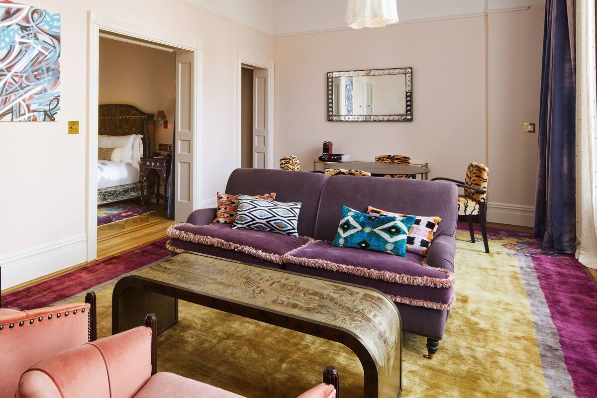 A well-furnished living room with a sofa, armchair, and a coffee table. There is a bed with a headboard in the background, and a mirror on the wall.