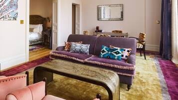 A well-furnished living room with a sofa, armchair, and a coffee table. There is a bed with a headboard in the background, and a mirror on the wall.