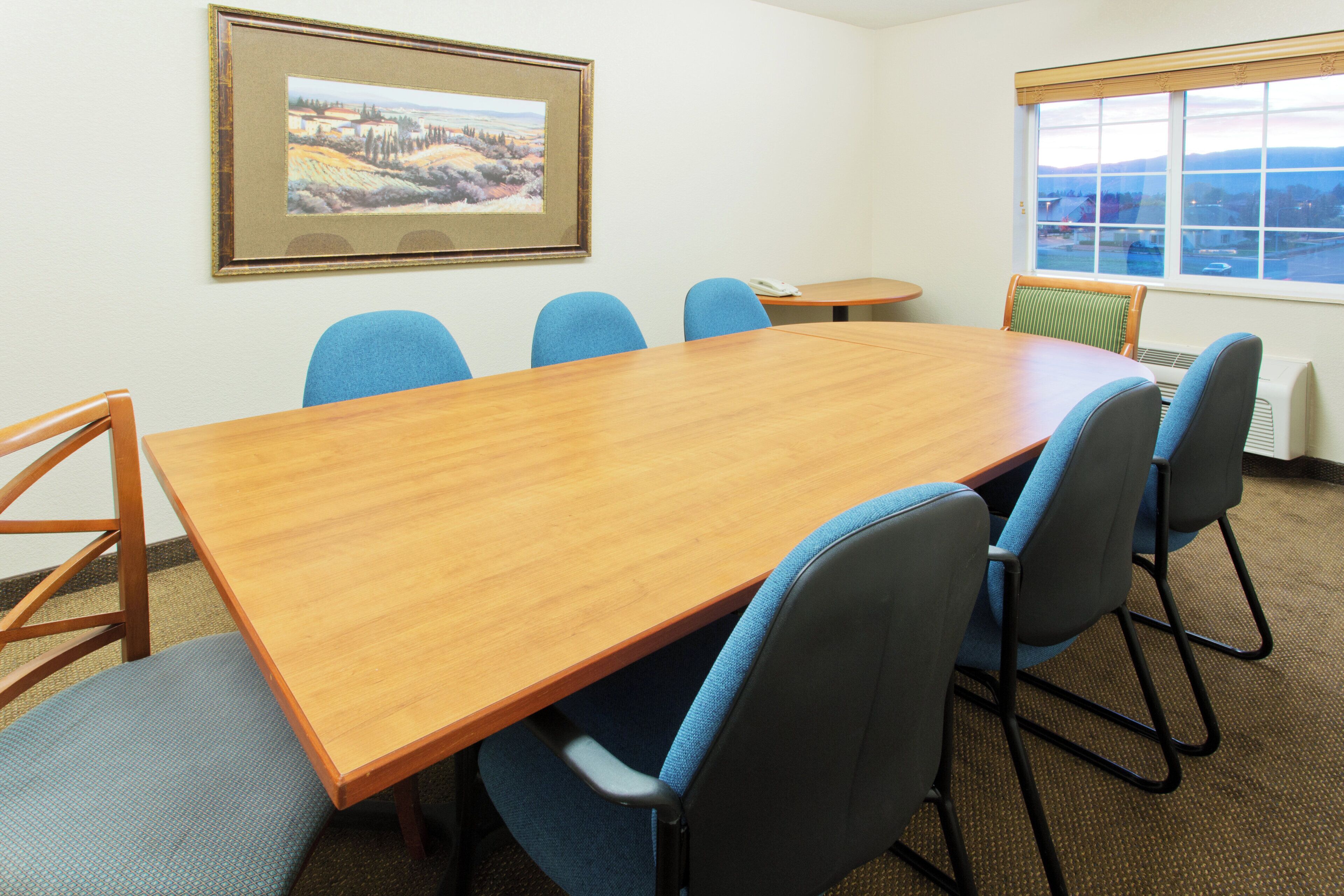 A conference room with a wooden table, blue chairs, a small round table, a telephone, and a window with a view.