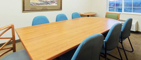 A conference room with a wooden table, blue chairs, a small round table, a telephone, and a window with a view.