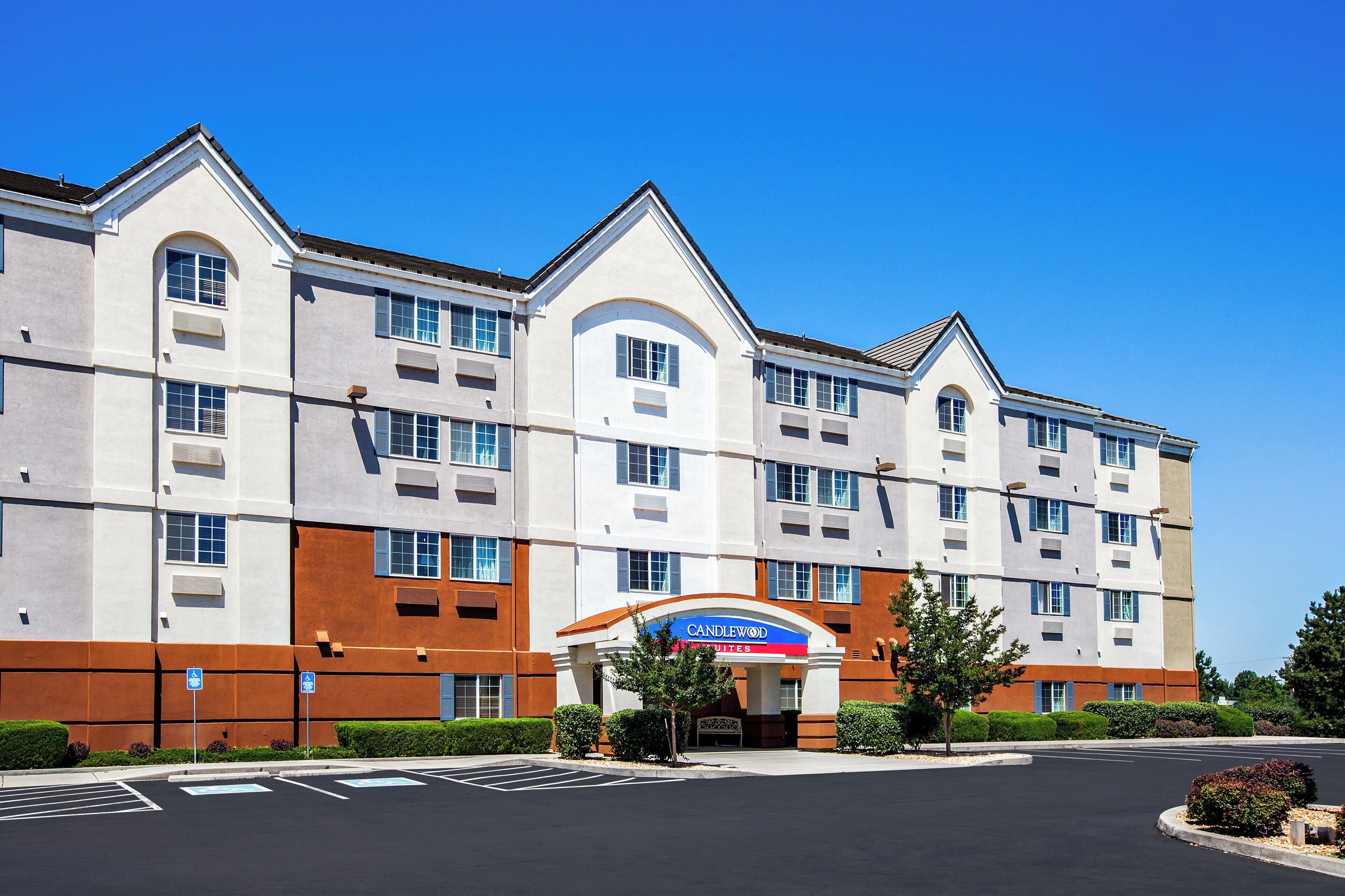 A multi-story hotel with a prominent 'Candlewood Suites' sign, a parking lot, and a clear blue sky.