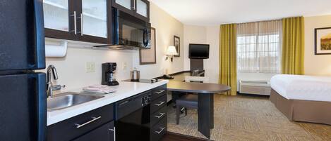 Una habitación de hotel compacta con kitchenette, una cama, un televisor y un ventanal con cortinas.