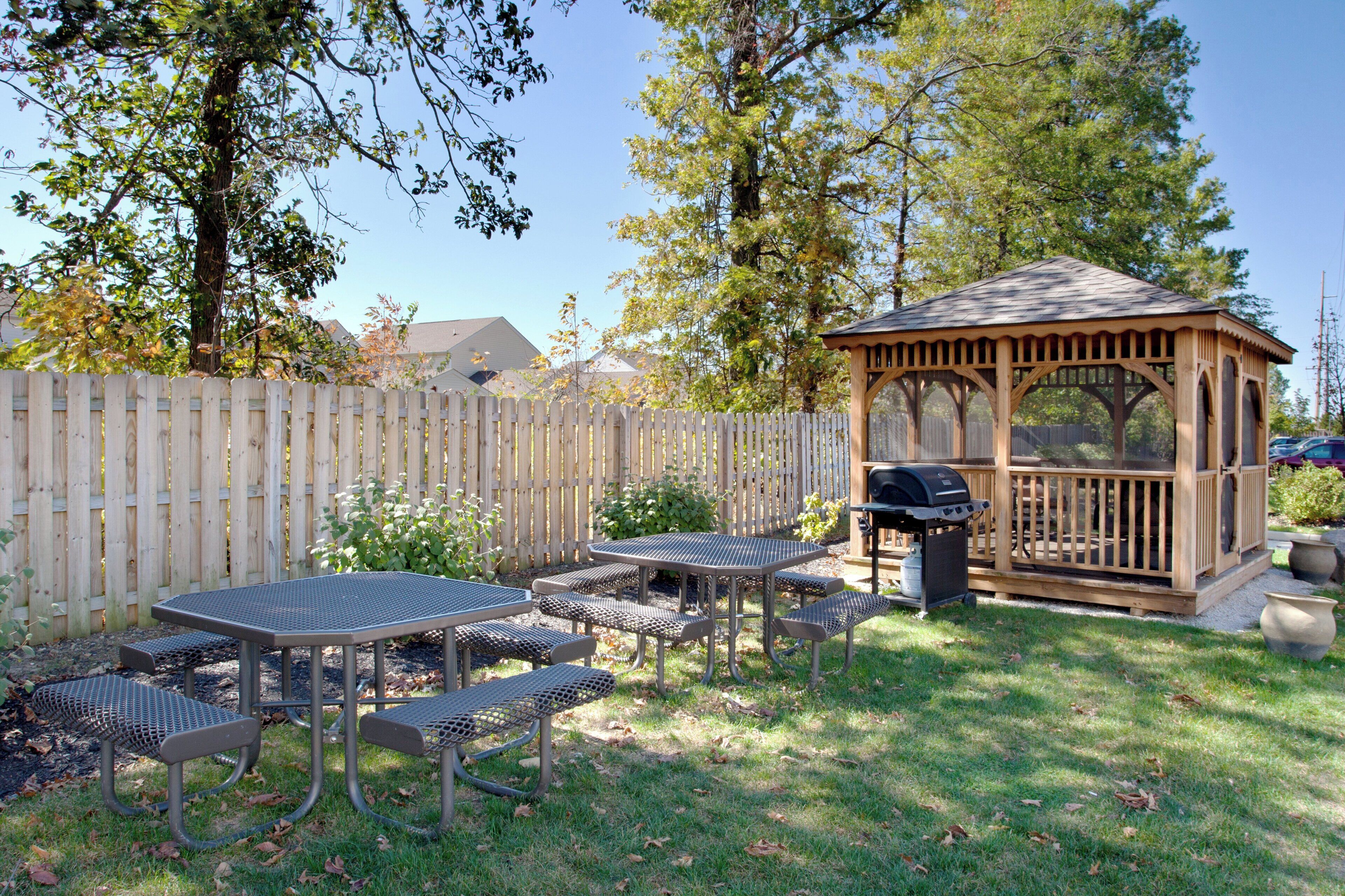 Un patio trasero con un gazebo de madera, una parrilla para barbacoa y un patio con mesas y bancos de metal.