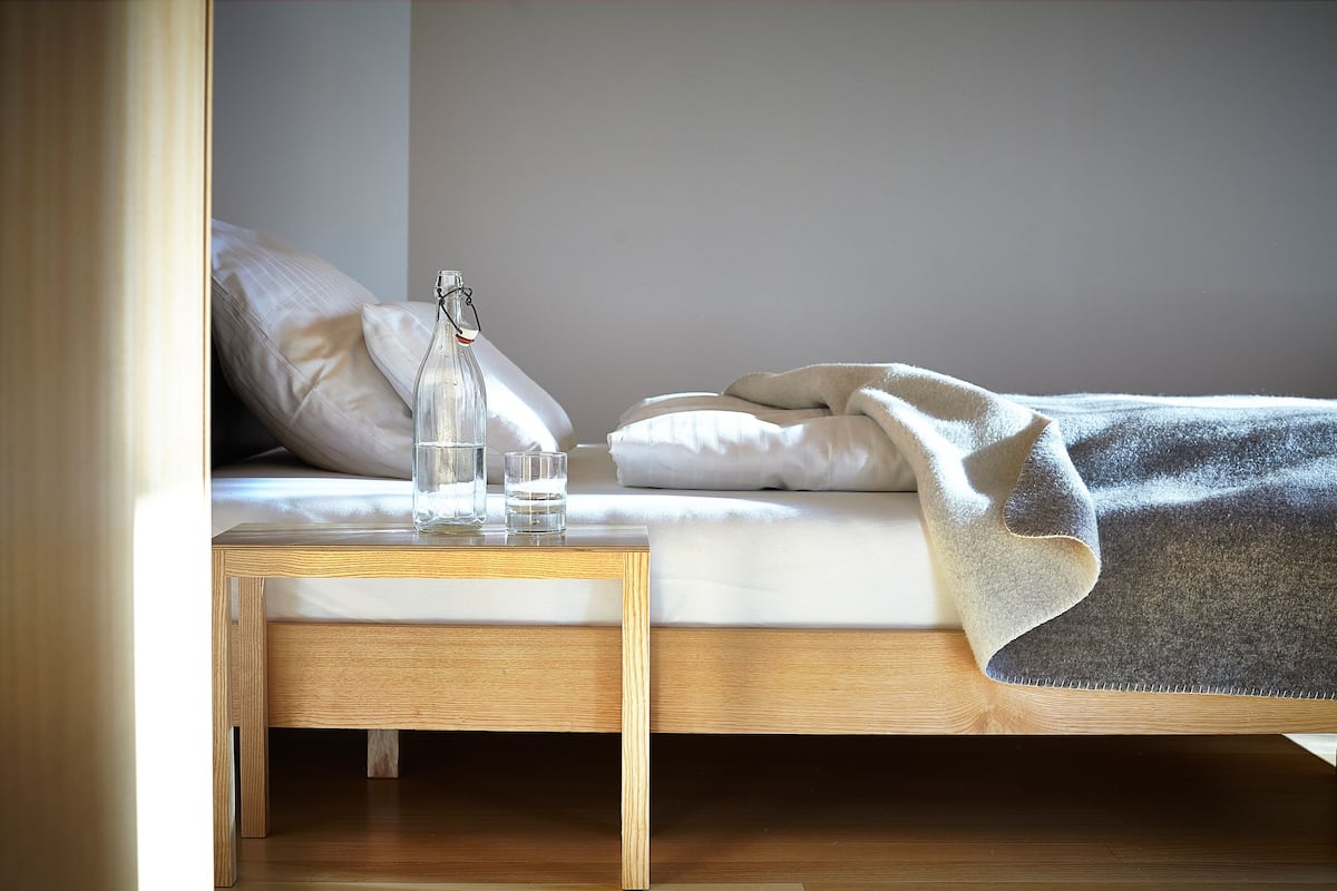 Una cama bien tendida con una almohada blanca, una manta gris y una mesita de noche de madera con una botella de vidrio y dos vasos.