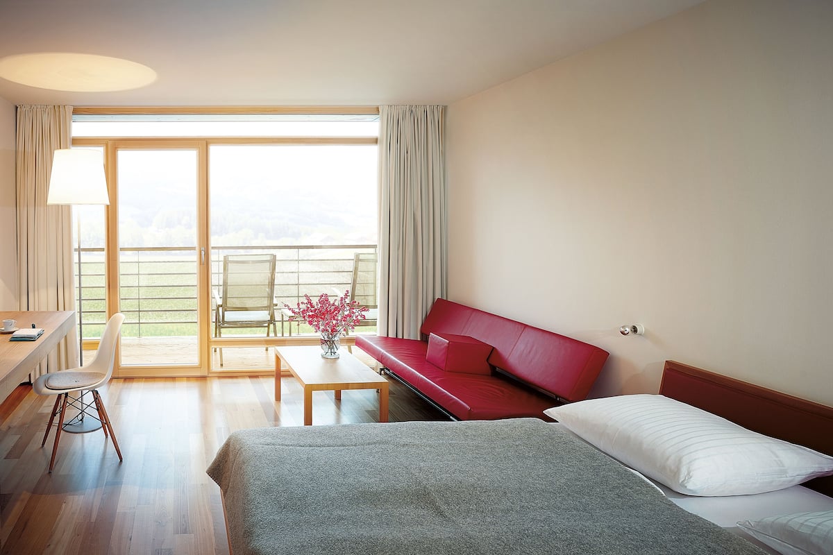 Un cuarto de hotel con una cama, un sofá rojo, una mesa de comedor, una silla y vistas al exterior.