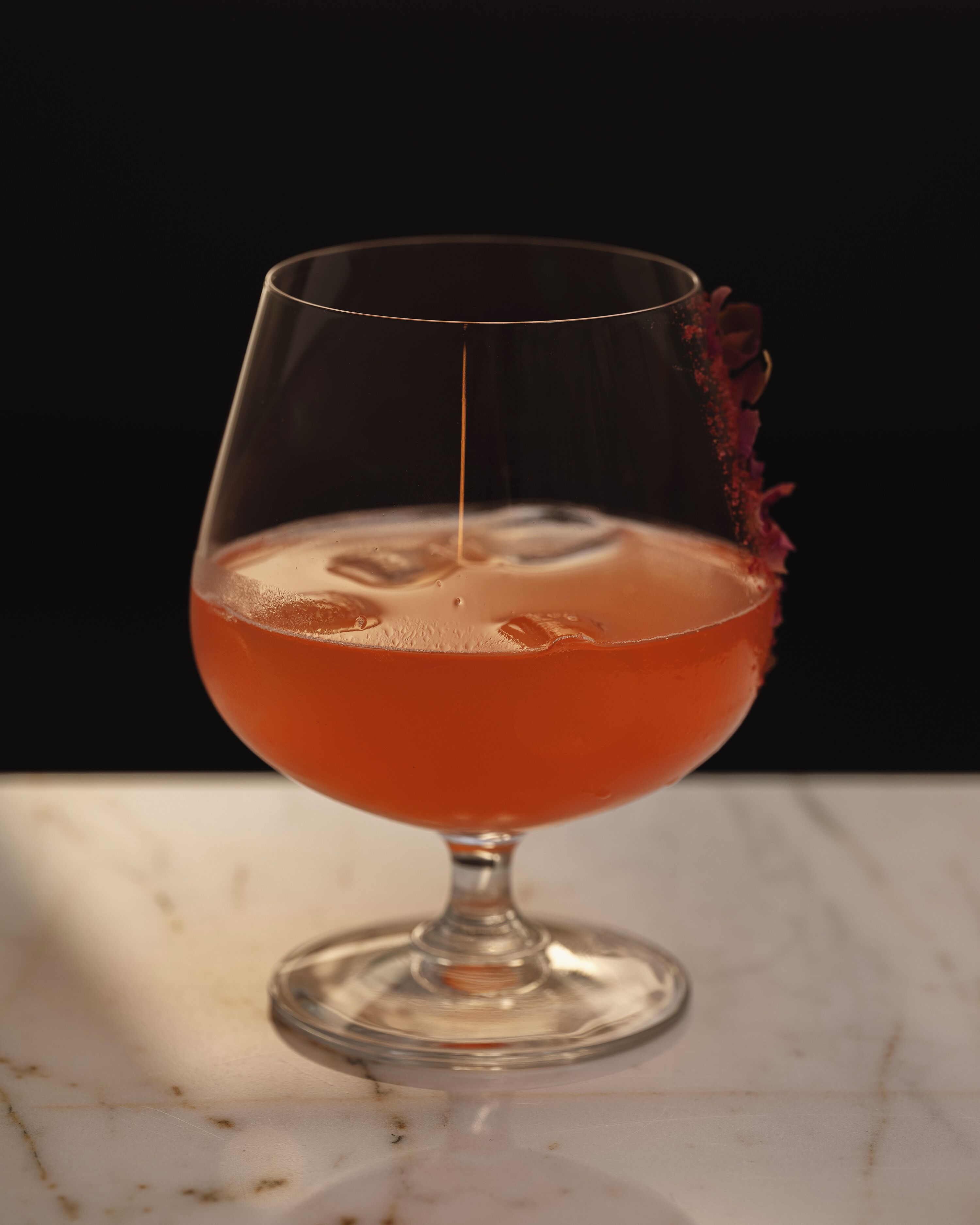 A clear glass with a stem and base, containing a red liquid, possibly a cocktail, with ice cubes.