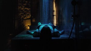 Una persona recostada sobre una camilla de masaje en una habitación poco iluminada, con pared de piedra y fuente de agua.