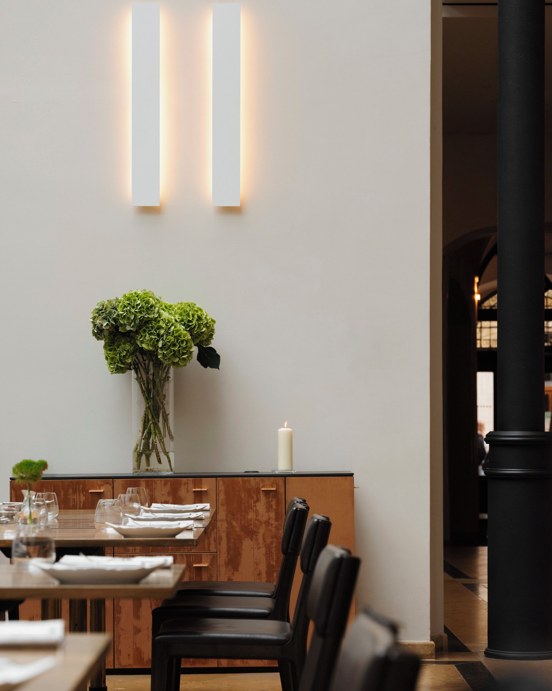 A dining area with a wooden table set for two, a vase of green foliage, and a candle on the table.