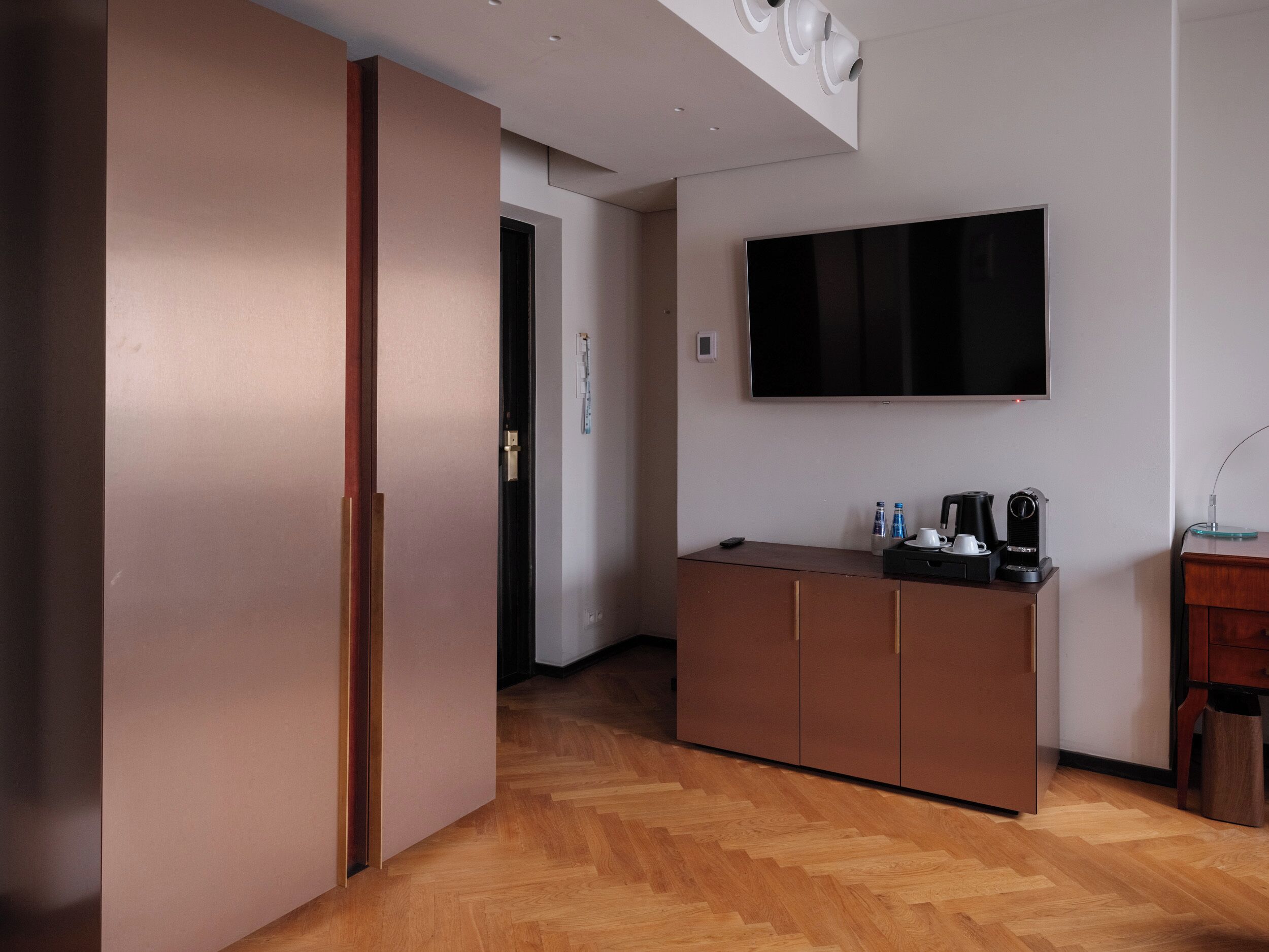 Ein modernes Hotelzimmer mit Holzboden im Fischgrätmuster, einem an der Wand montierten Flachbildfernseher, einer Minibar und einem Holzkabinett.
