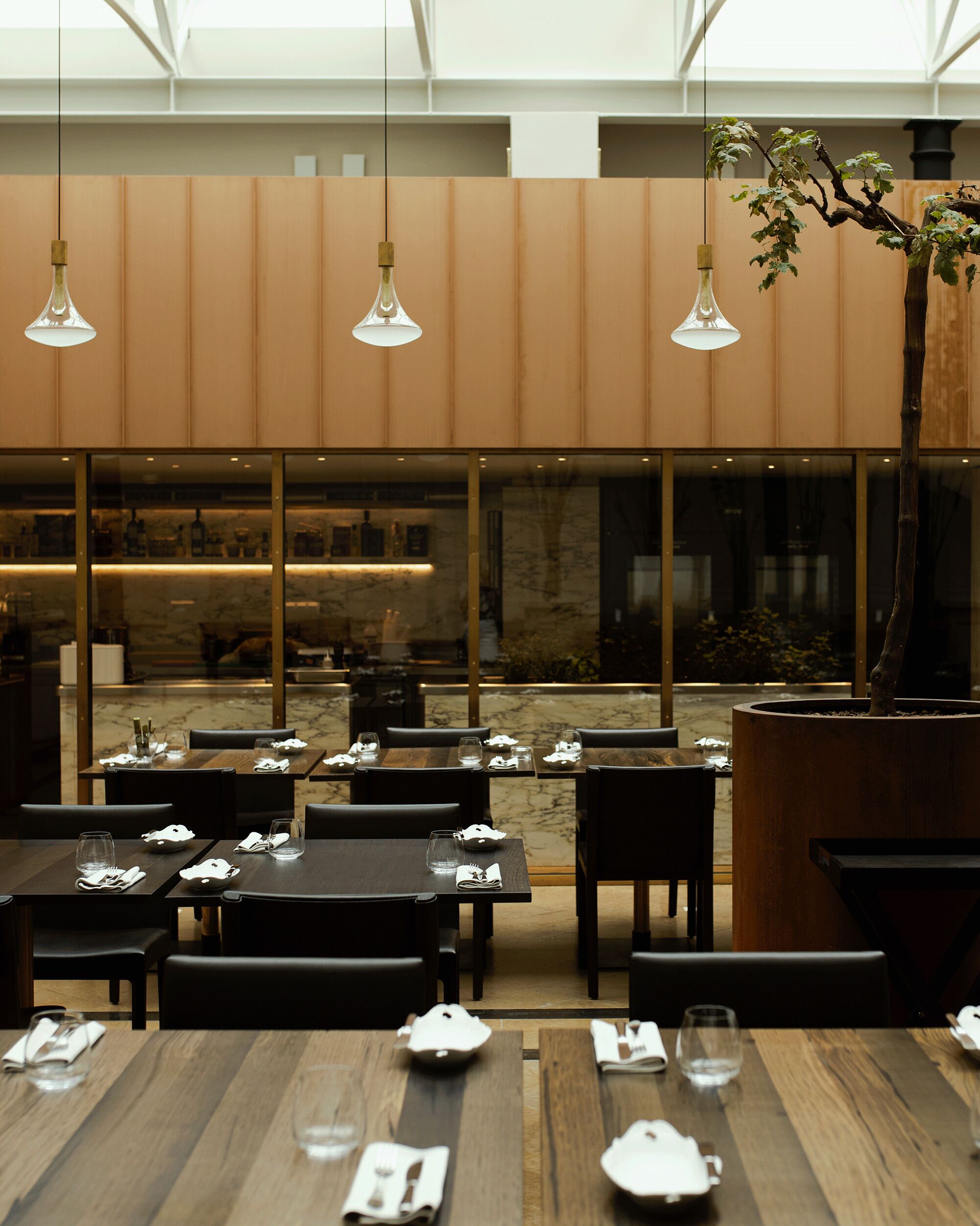 A restaurant with wooden tables and chairs, hanging pendant lights, and a large window overlooking a garden.