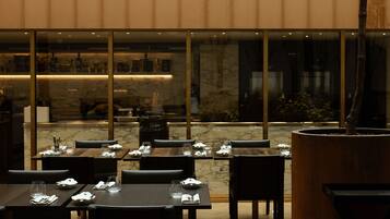 A restaurant with wooden tables and chairs, hanging pendant lights, and a large window overlooking a garden.
