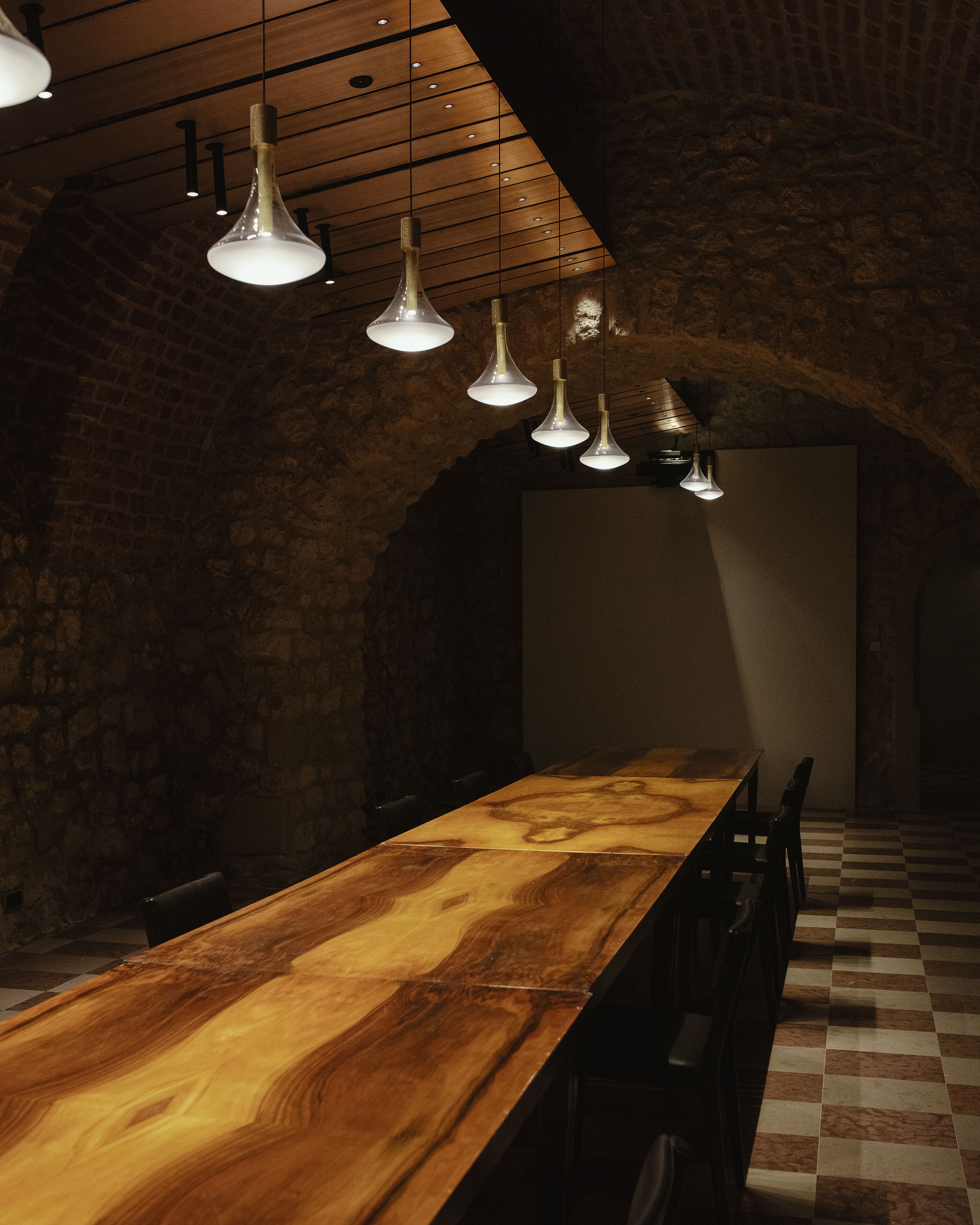 Ein langer Holztisch mit Stühlen, hängende Pendelleuchten und eine Steinwand.