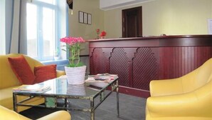 A reception area with a reception desk, a glass-top table, yellow chairs, a potted plant, and a window with curtains. - Sesame (Riga)