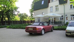 A red convertible parked in a paved driveway with a white car beside it. - Sesame (Riga)