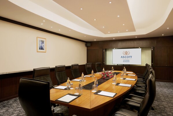 Una sala de conferencias con una larga mesa, sillas, botellas de agua y una pantalla de proyector mostrando 'ASCOTT SATHORN BANGKOK'.