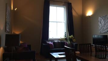 Een woonkamer met een bank, een salontafel en een eettafel met stoelen. Er is een raam met gordijnen, een aan de muur bevestigde televisie en een schilderij.