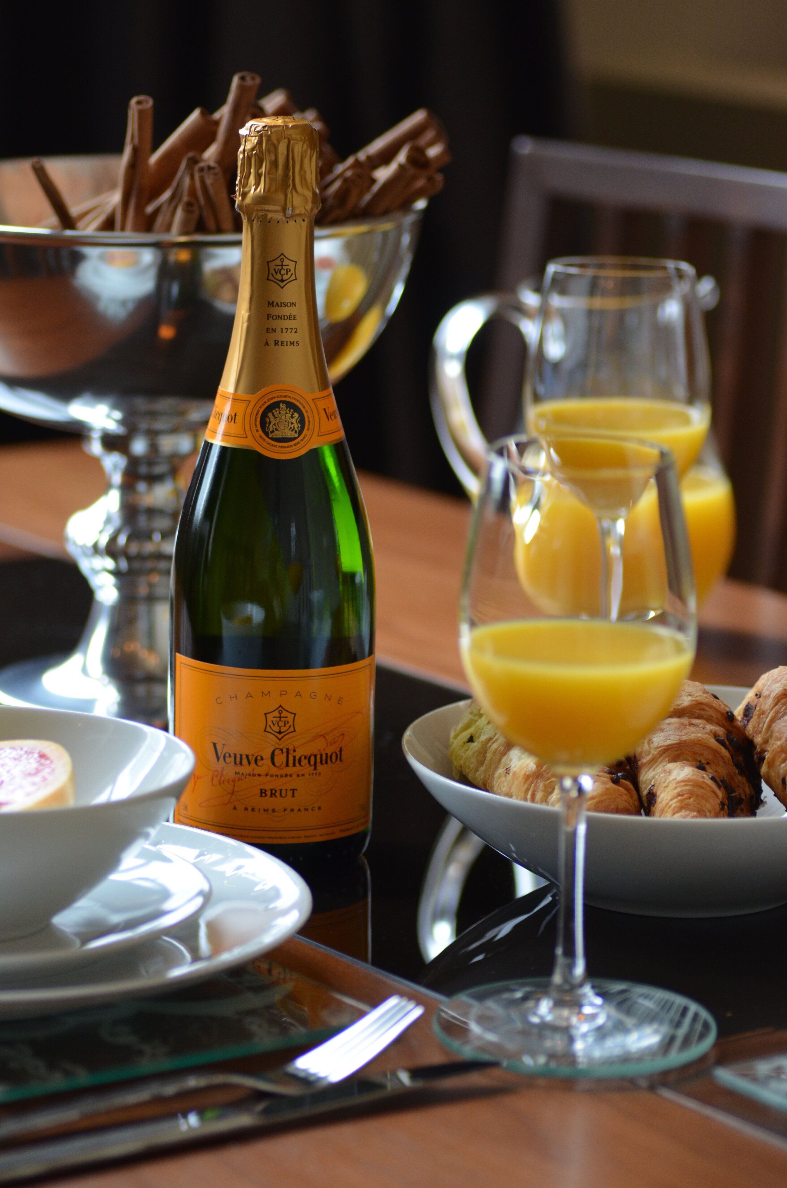 Une bouteille de champagne Veuve Clicquot, un panier de croissants et une carafe de jus d’orange sur une table.