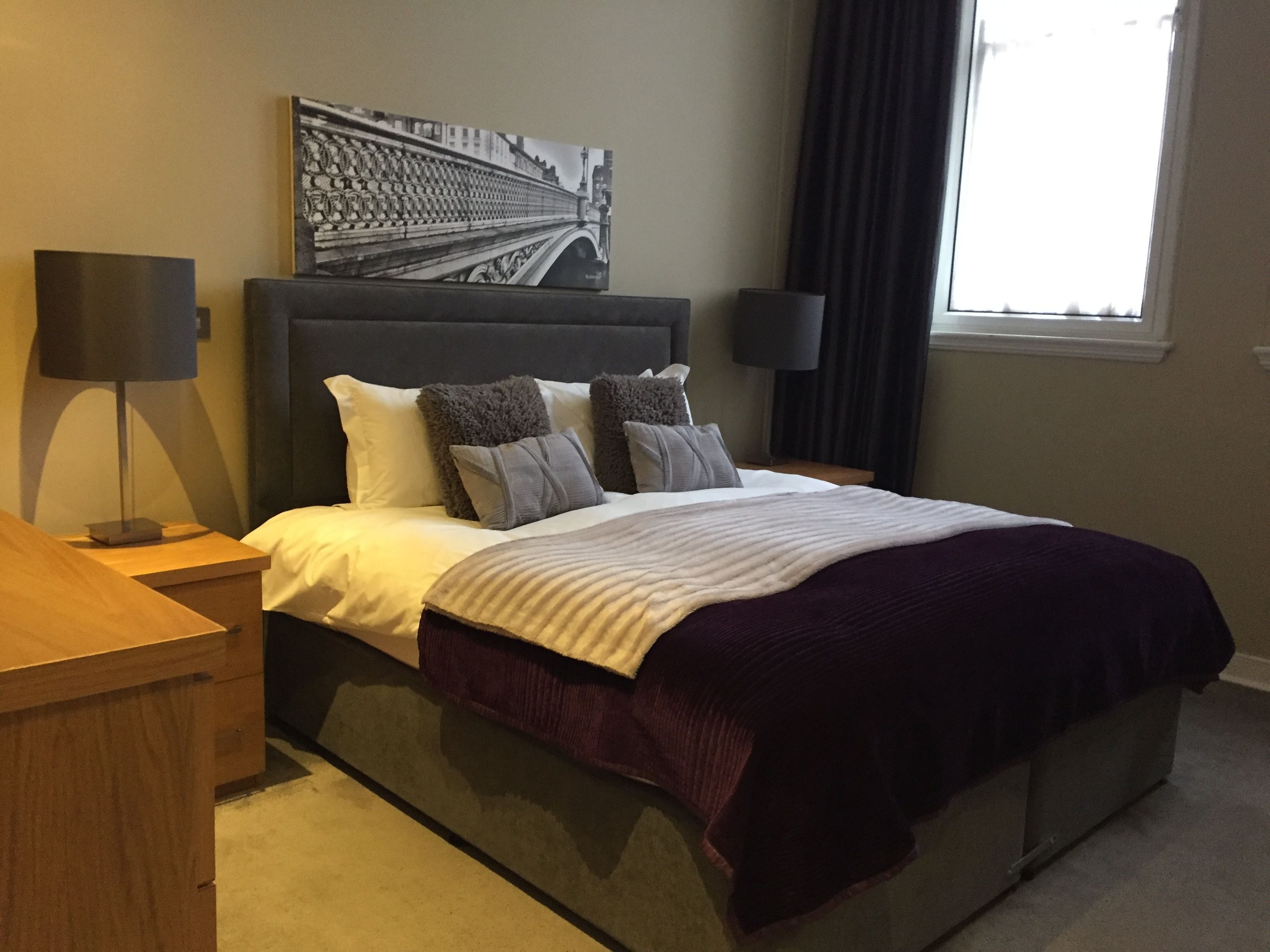 A hotel room with a large bed, a headboard, two bedside tables, a lamp, a window with curtains, and a wall-mounted artwork.